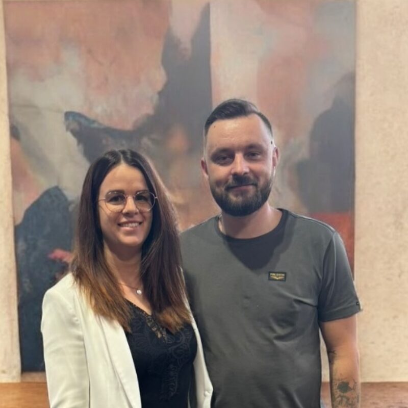 Lisa ist eine Frau mit langen braunen Haaren und Brille steht neben einem bärtigen Mann Robin in einem grauen T-Shirt; beide lächeln vor einem verschwommenen abstrakten Gemälde.