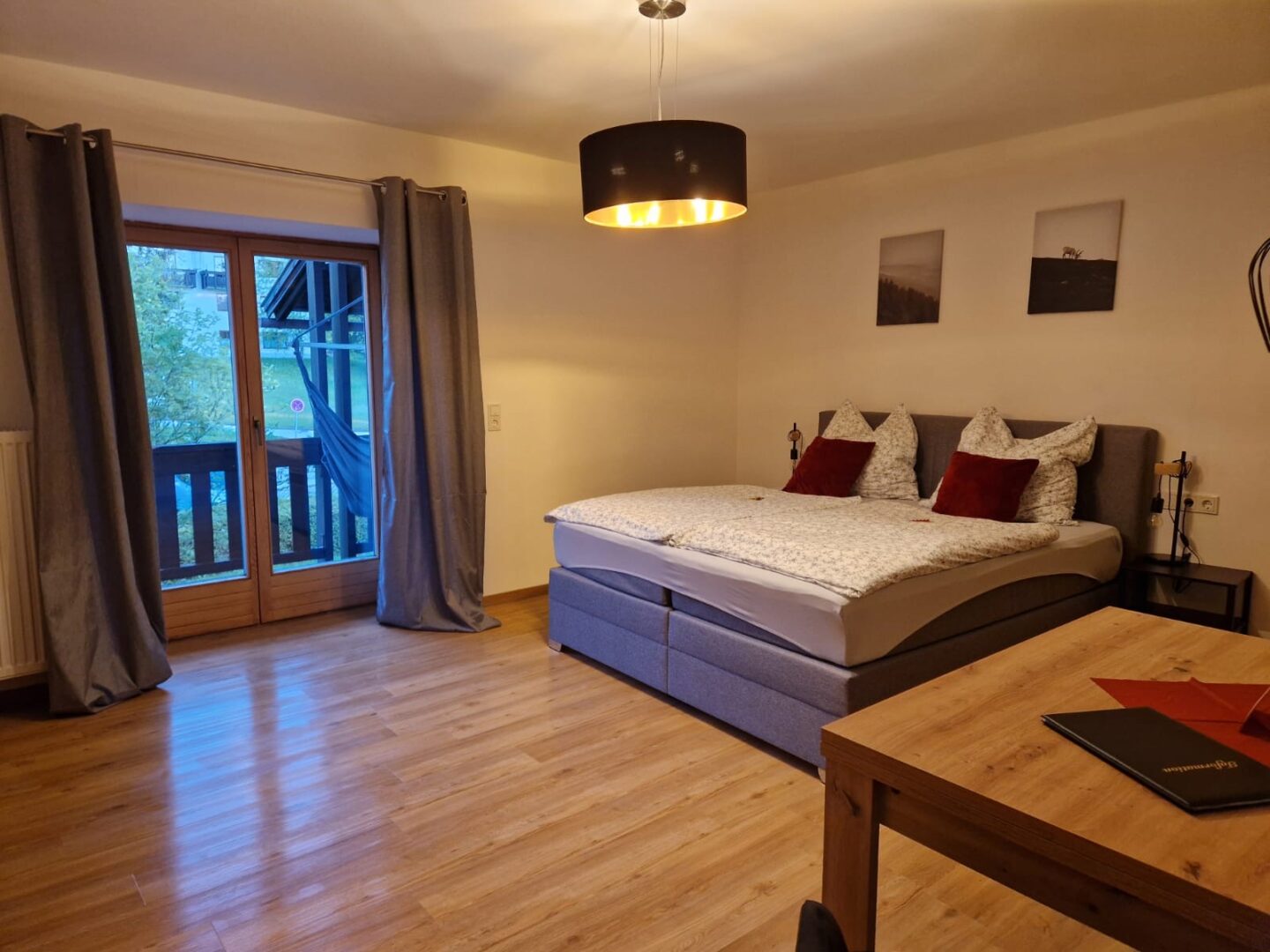Ein gemütliches Schlafzimmer mit Doppelbett, weiß-grauer Bettwäsche, roten Kissen, Holzboden und Holztisch. Große Glastüren mit grauen Vorhängen führen auf einen Balkon mit Hängematte. Warmes Deckenlicht.