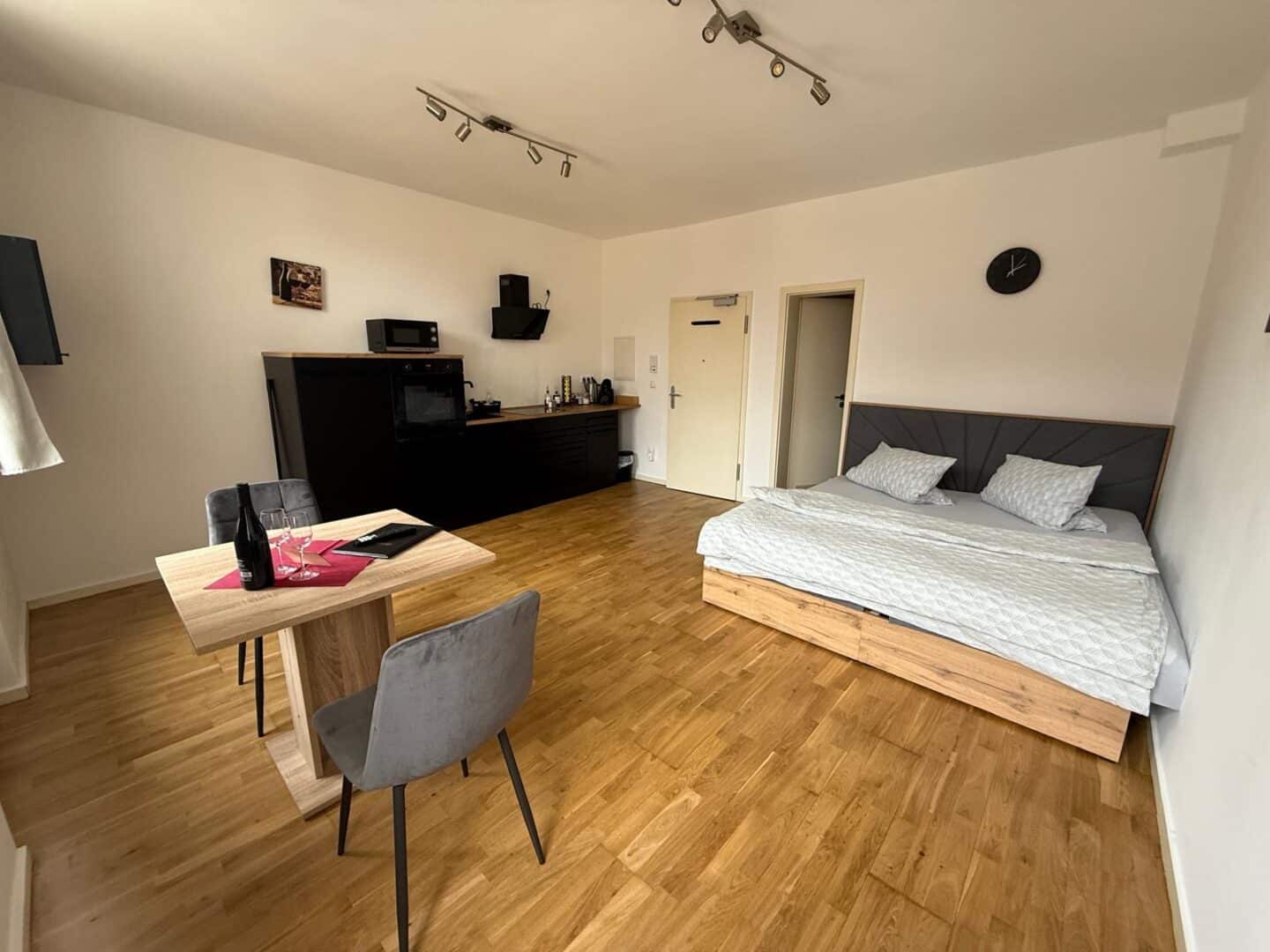 Ein modernes Studio-Apartment mit Holzboden, Doppelbett, kleinem Esstisch mit zwei Stühlen, kompaktem Küchenbereich, Wanduhr und minimalistischem Dekor, alles in neutralen Tönen.