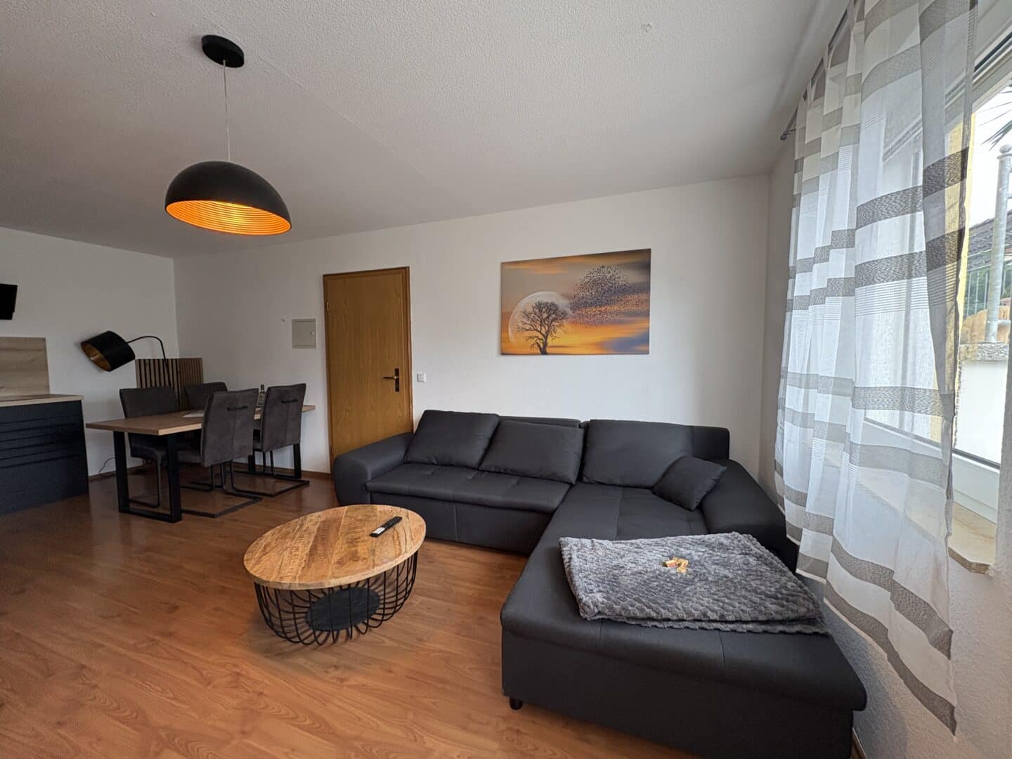Ein modernes Wohnzimmer mit einem schwarzen Sofa, einem Couchtisch aus Holz und einem Essbereich mit Tisch und vier Stühlen. Große Fenster mit transparenten Vorhängen lassen Tageslicht herein; an der Wand hängt ein Landschaftsgemälde.