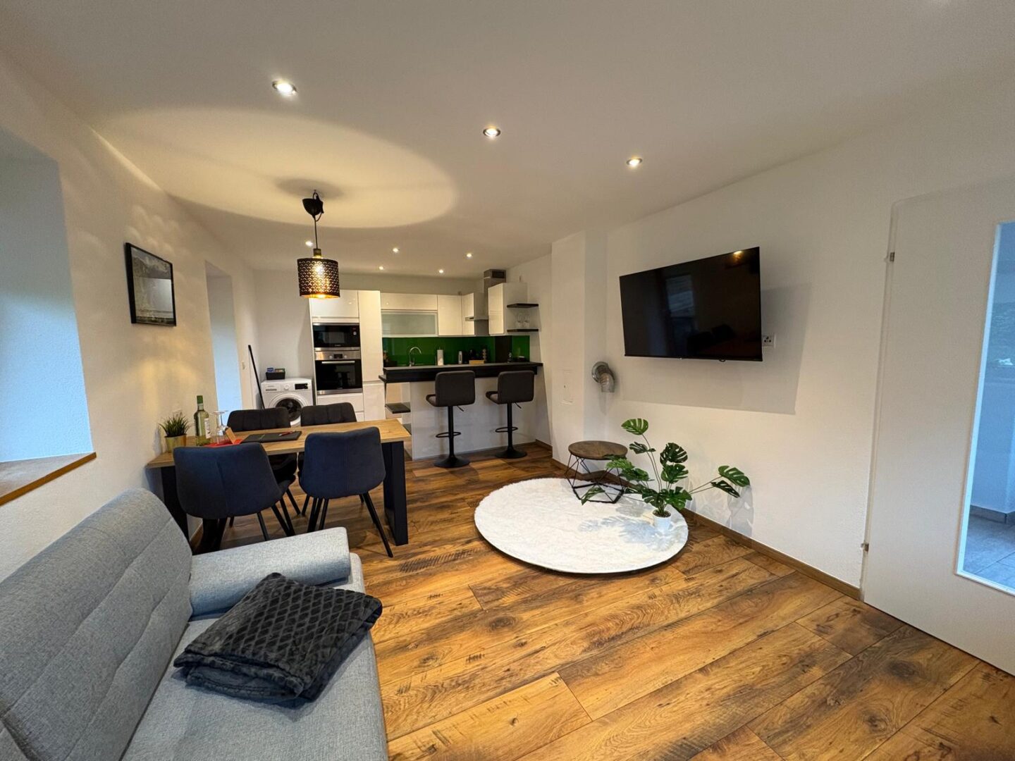 Moderner offener Wohnraum mit Holzfußboden, grauem Sofa, Esstisch mit dunklen Stühlen, Küche mit weißen Schränken und grüner Aufkantung, an der Wand montiertem Fernseher, kleinem runden Tisch und einer Topfpflanze.